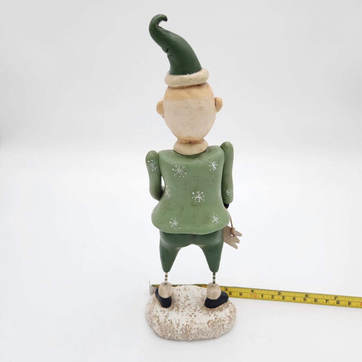 A hand-painted Christmas  Janell Berryman Pumpkinseeds "Sage and Peppermint Santa shown from the back, standing on a textured base with a yellow tape measure placed alongside to indicate its height. The gnome is wearing a green pointed hat and a green patterned sweater, with a beige face and hands.