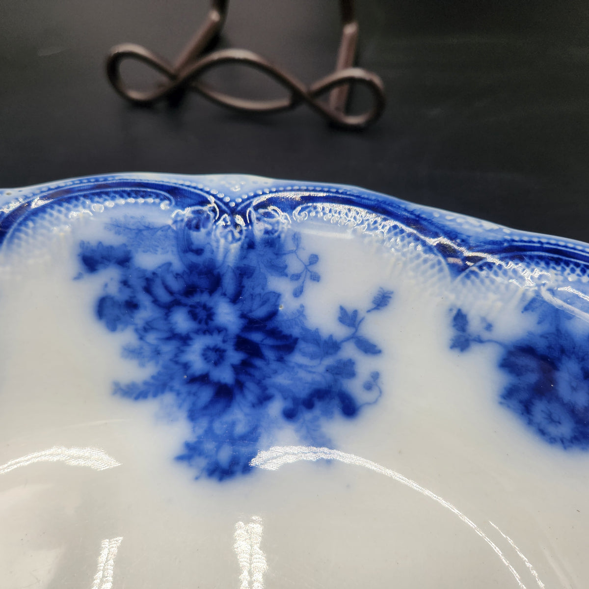 Close-up of a white ceramic plate with blue floral patterns and a scalloped rim, photographed against a dark background with part of a metal stand visible. Antique 1890s Flow Blue Cobalt Oval Serving Bowl Deep Vegetable Dish