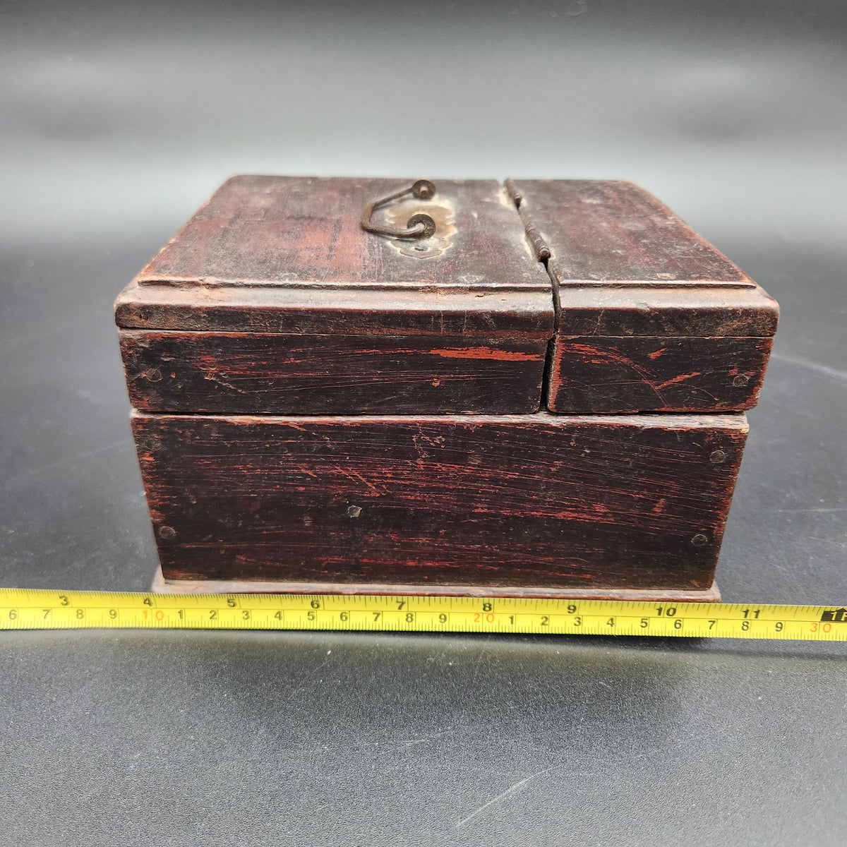 A small rectangular dark reddish-brown wooden box with a metal latch on the front, shown next to a yellow tape measure on a gray surface.