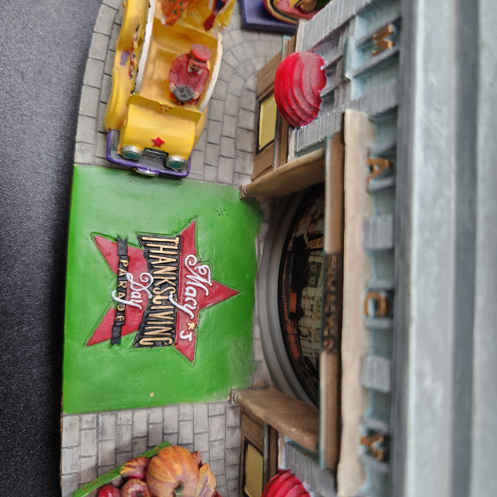 A close-up, angled view of a miniature diorama or toy model set, featuring a green sign with a red star and the text 'Halloween' and 'Thanksgiving' alongside a yellow toy car and a gray building structure. Santa snowglobe Macy's Day Parade 2003