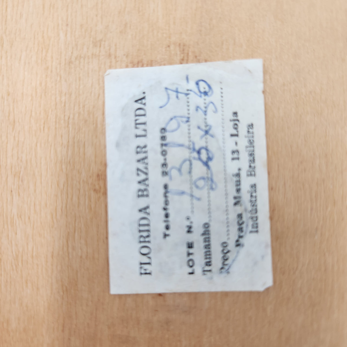 A small rectangular printed paper label with Portuguese text and handwritten blue ink markings, placed on a light wood surface. The label includes the brand name 'FLORIDA BAZAR LTDA. Vintage Brazilian Butterfly Wing Marquetry Tray Rio De Janeiro Inlaid Wood Label
