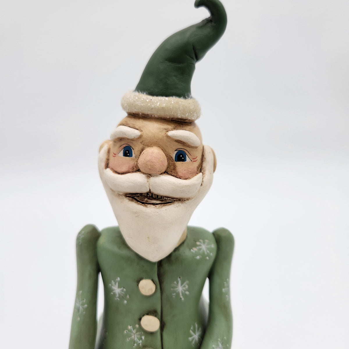 A handmade  Janell Berryman Pumpkinseeds "Sage and Peppermint Santa of a whimsical elf or gnome with a green hat and matching green jacket decorated with white snowflake patterns, set against a plain light gray background.