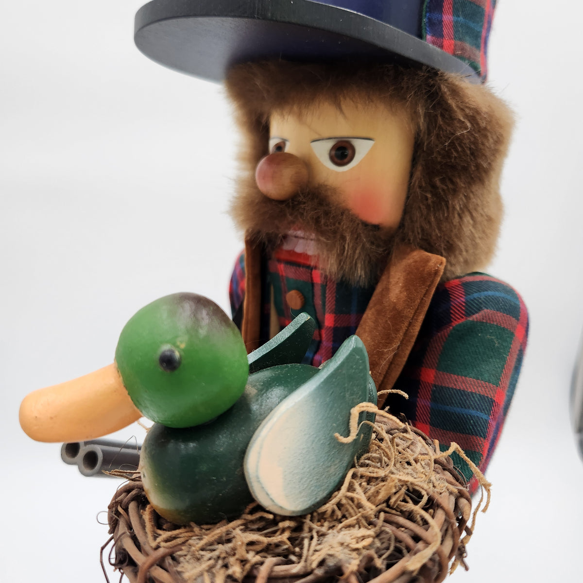 A rustic  Christian Ulbricht Nutcracker The Duck Hunter wearing a plaid hat and jacket with a brown beard and hat fur, holding a green and white wooden duck in a nest on a white background.
