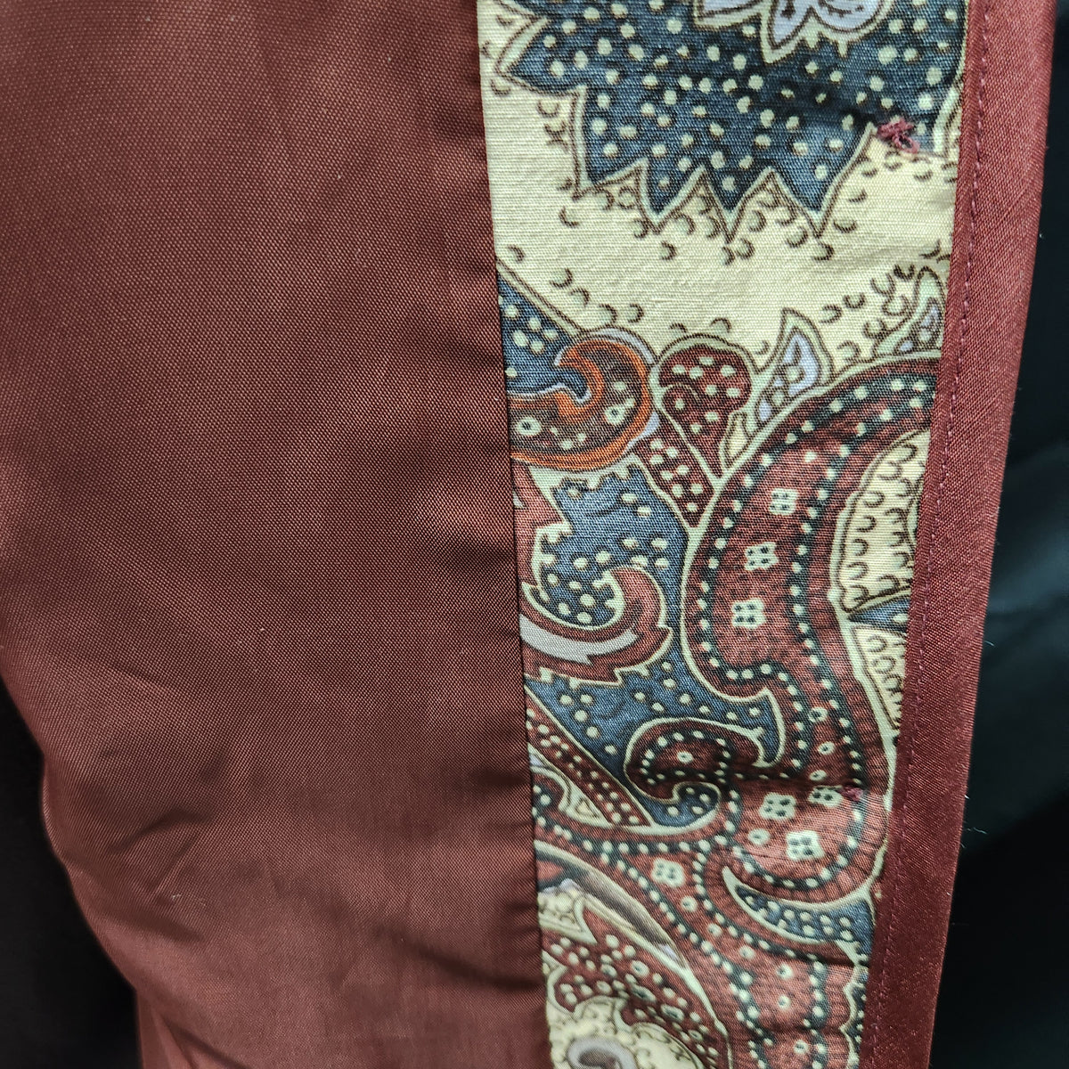 Close-up of a silk garment showing a solid rust-brown fabric panel next to a vertical paisley patterned trim in cream, blue, and rust-brown.