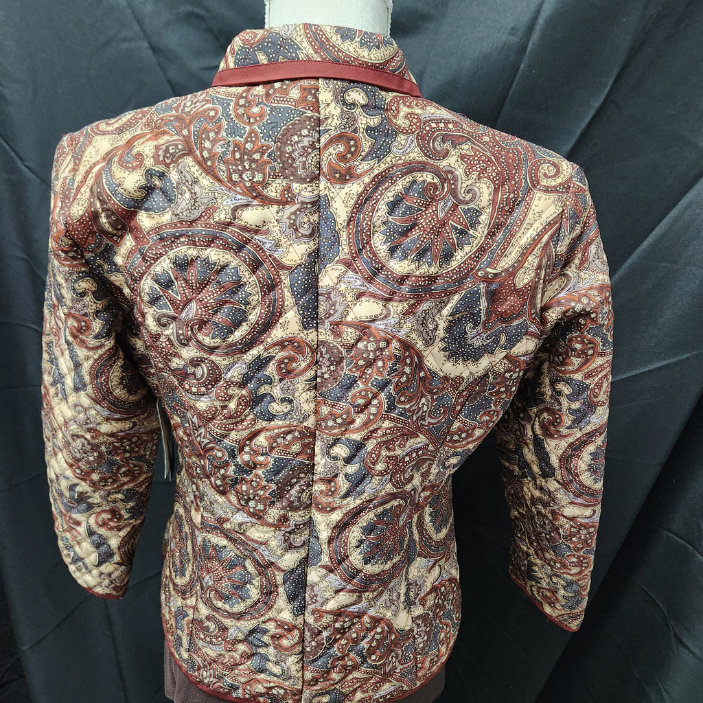 A Silk quilted jacket with an intricate paisley pattern in beige, brown, and blue, displayed on a mannequin against a dark gray fabric backdrop.