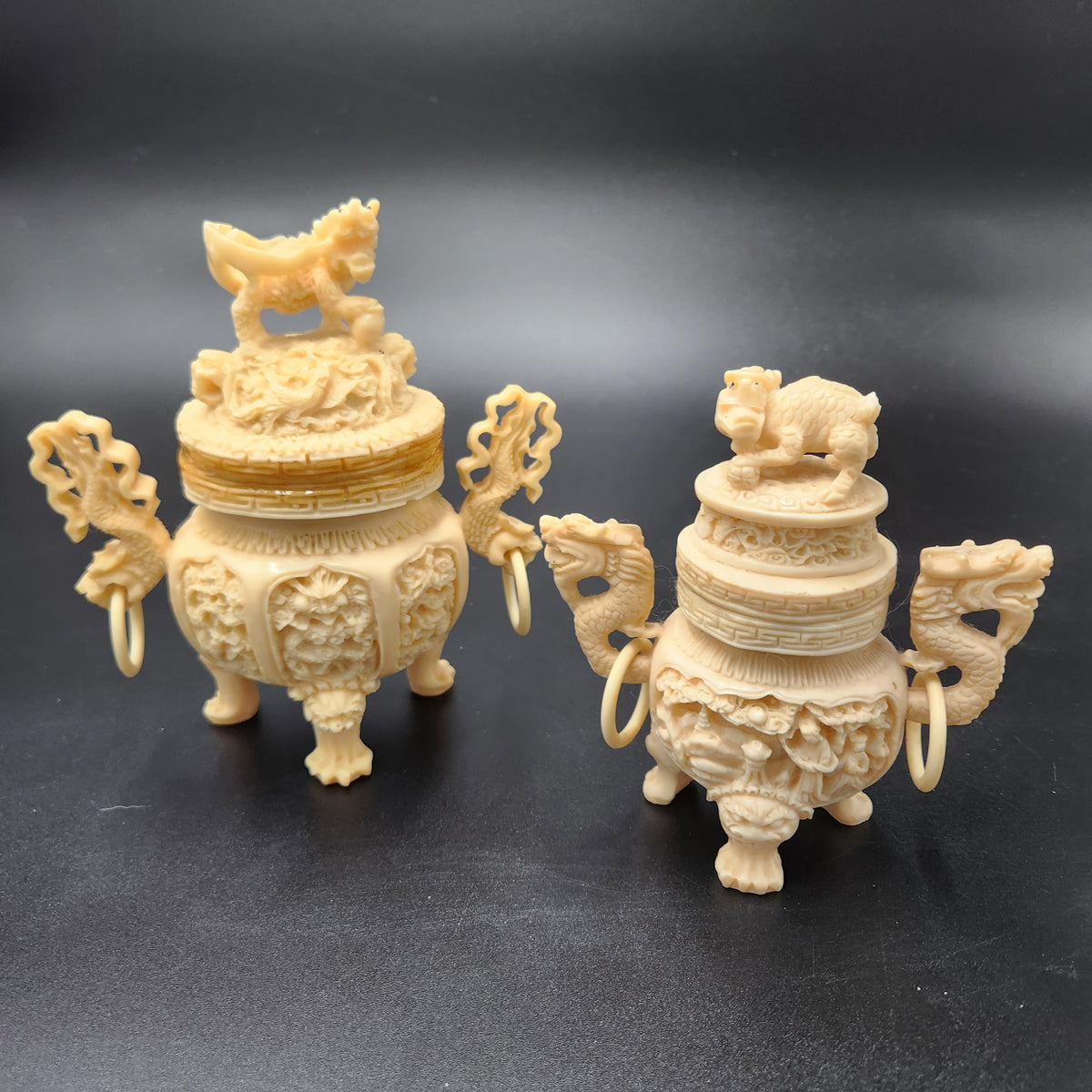 Two ornate cream-colored carved figurines resembling dragon-shaped censer pots with animal toppers on the lids, displayed on a dark gray background. Vintage Chinese Carved Resin Faux Ivory Incense Burners Tripod Censers 