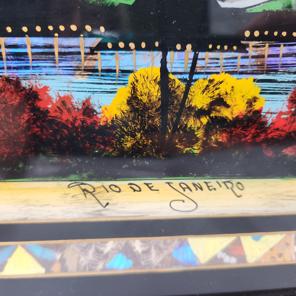 A close-up of a colorful artwork depicting trees and foliage in red, yellow, and blue tones, with the text 'Rio de Janeiro' written across a light wood plaque in front. Vintage Brazilian Butterfly Wing Marquetry Tray Rio De Janeiro Inlaid Wood Label