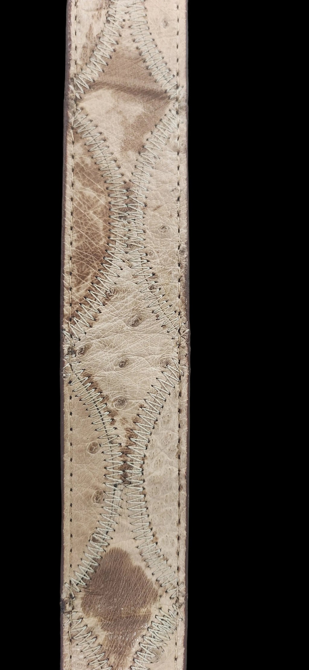 A close-up of a natural ostrich leather strap, showing its textured, mottled beige and brown skin pattern with whipstitched diamond-shaped overlays, set against a plain gray background.Justin Genuine Ostrich Belt