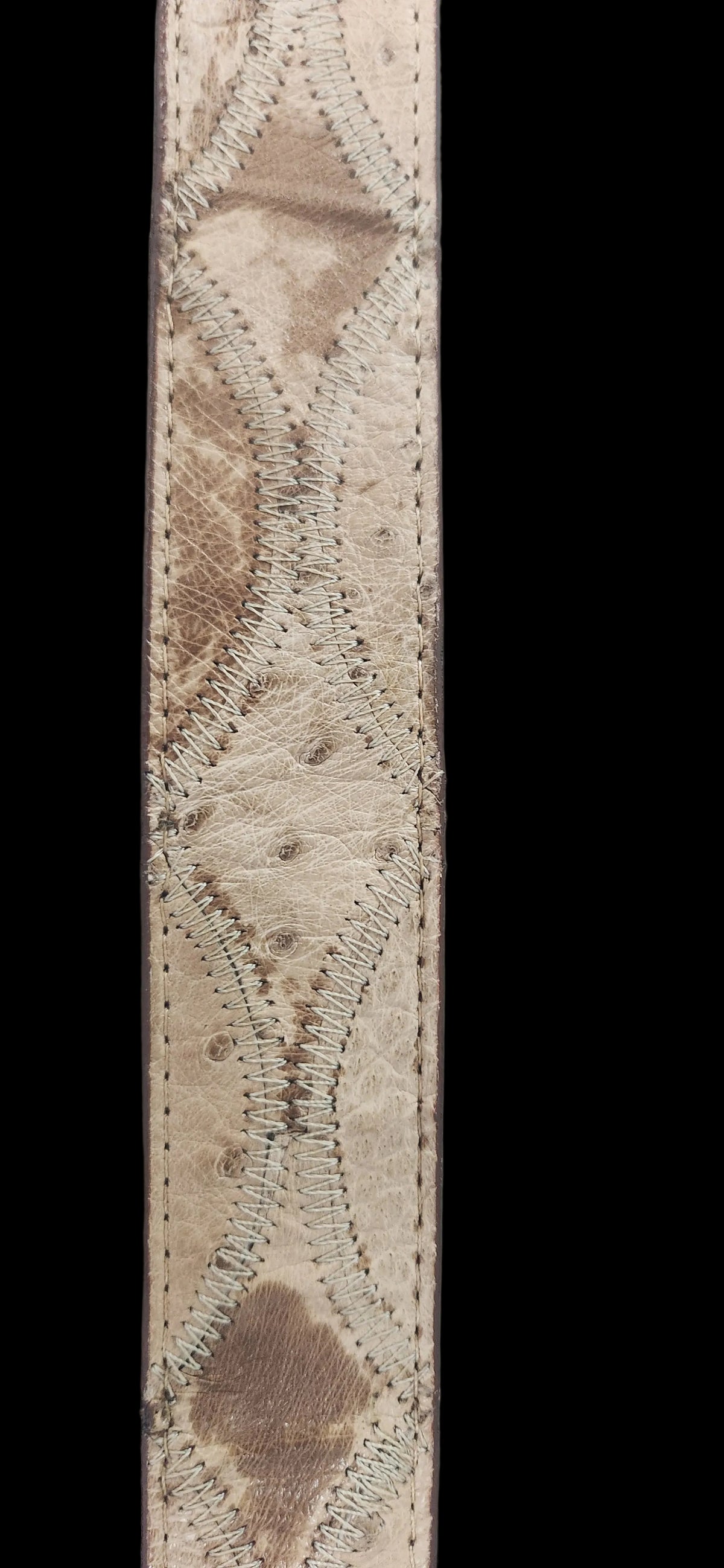 A close-up of a natural ostrich leather strap, showing its textured, mottled beige and brown skin pattern with whipstitched diamond-shaped overlays, set against a plain gray background.Justin Genuine Ostrich Belt