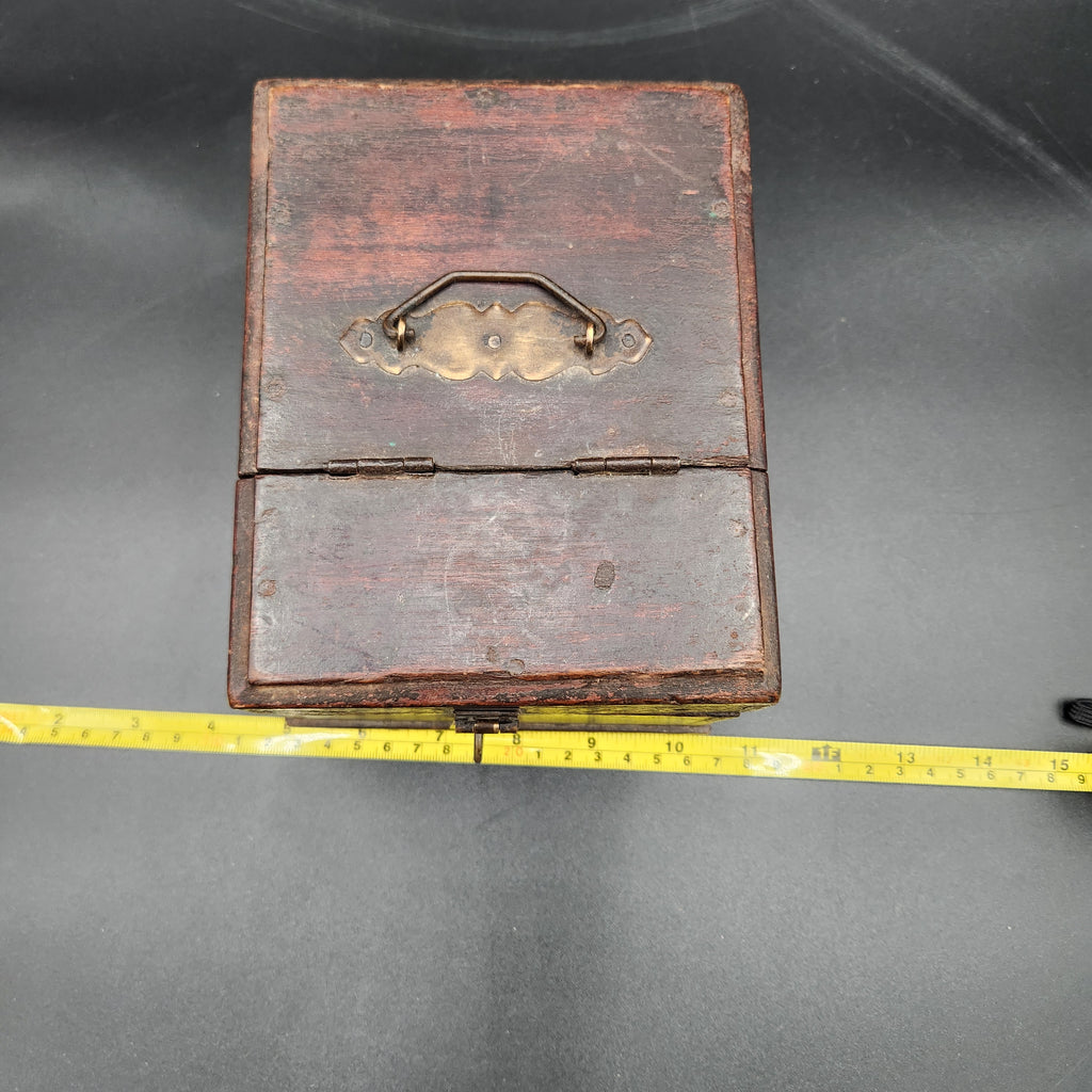 A small vintage wooden box with a hinged lid and brass handle, shown on a dark gray surface next to a yellow measuring tape for scale.