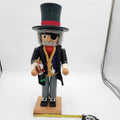 A painted wooden nutcracker figurine by Christian Ulbrickt  wearing a black top hat with a red band, black coat, gray pants, and a gold chain necklace, standing on a wooden base with a tape measure in front on a white studio background.