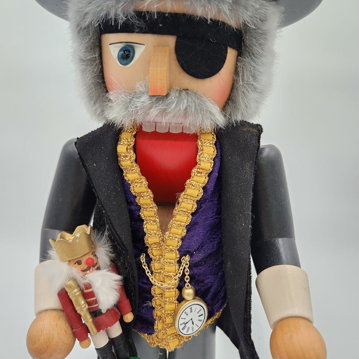 A close-up of a decorative nutcracker figure dressed as a pirate, with a gray furry hat and beard, black eyepatch, black jacket, purple vest, gold chain necklace with a pocket watch pendant, and a small miniature pirate figure in one hand, against a light gray background. Christian Ulbrickt