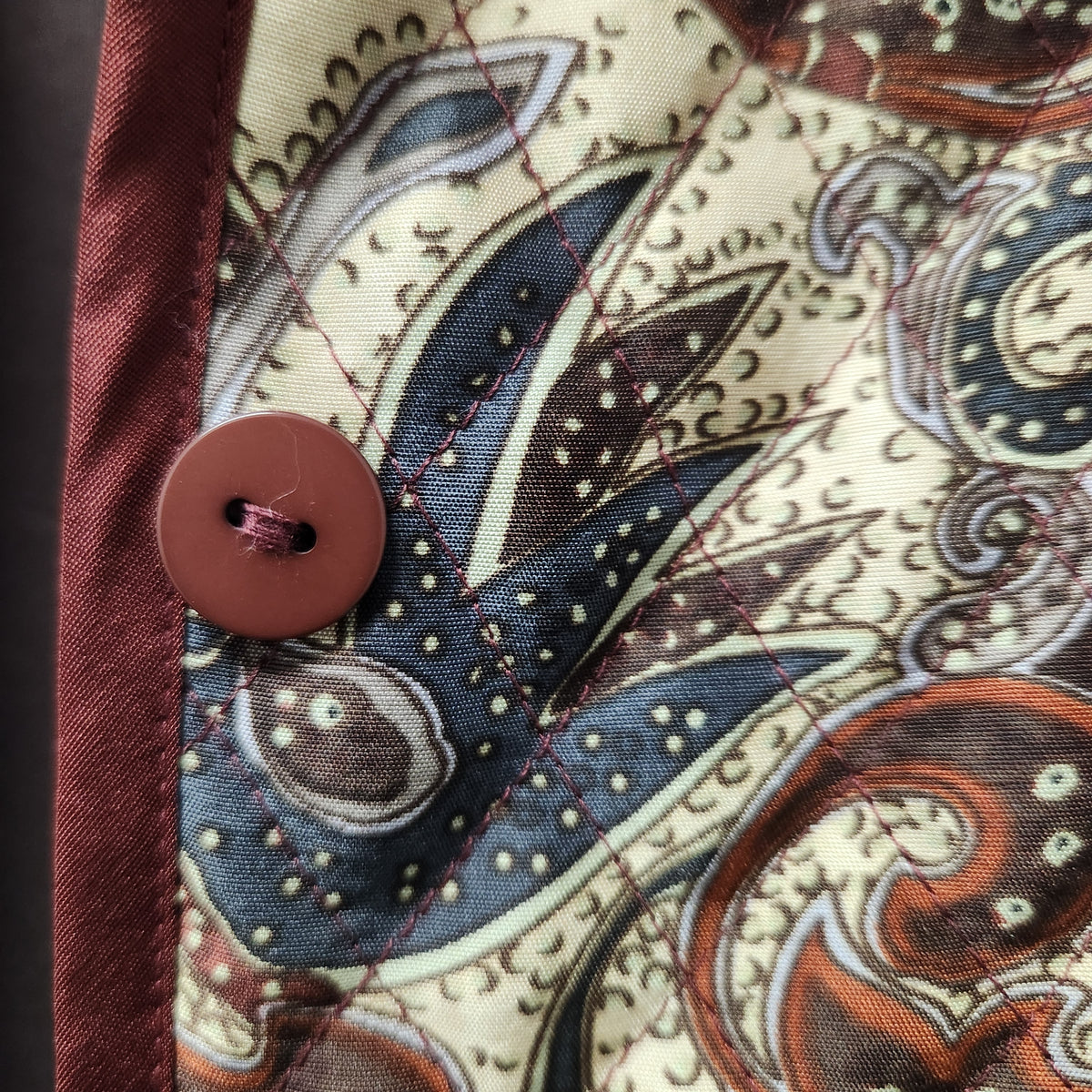 Close-up of a patterned fabric item with a paisley print in cream, blue, brown, and rust tones, featuring a brown button and a dark red stitched border.
