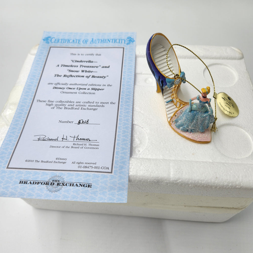An ornament displayed on a white foam insert next to an authenticity certificate from The Bradford Exchange. The ornament is shaped like a high-heeled slipper in blue and gold, with a small figure of a woman in a blue dress inside.