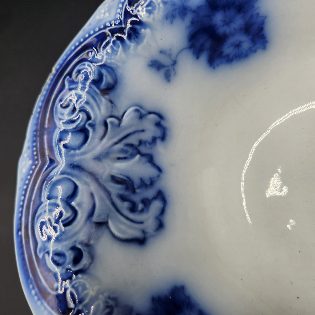 Close-up of a blue and white porcelain plate showing an embossed floral pattern along the rim and a glossy white interior glaze. Antique 1890s Flow Blue Cobalt Oval Serving Bowl Deep Vegetable Dish