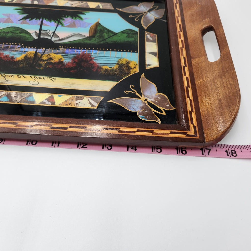 A close-up of a wooden serving tray with a colorful landscape illustration and butterfly inlay, shown next to a pink measuring tape to indicate size. Vintage Brazilian Butterfly Wing Marquetry Tray Rio De Janeiro Inlaid Wood Label