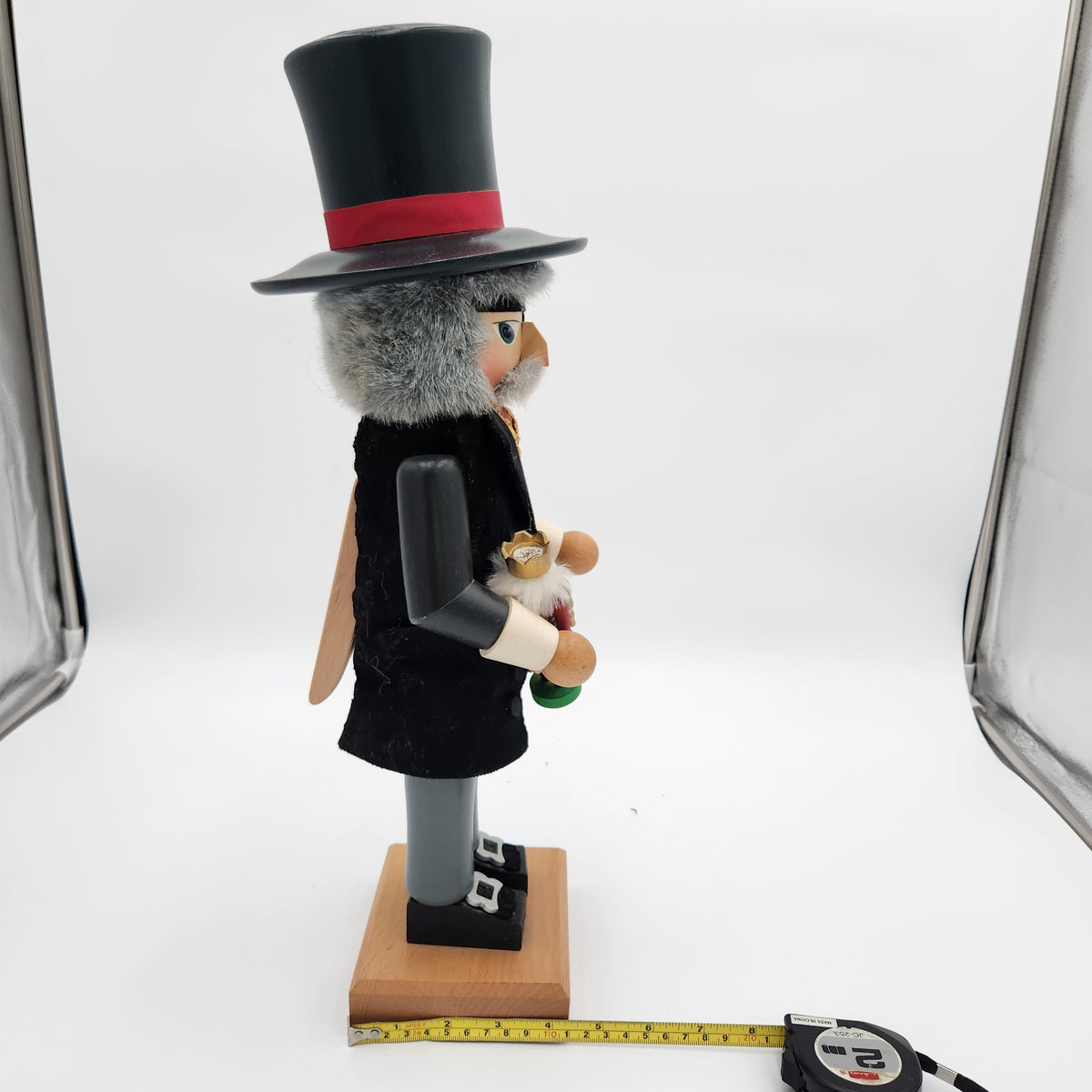 A handcrafted nutcracker figurine of a bird character in a black top hat with a red band, black coat, gray legs, and a wooden base, shown with a tape measure on a white studio background.