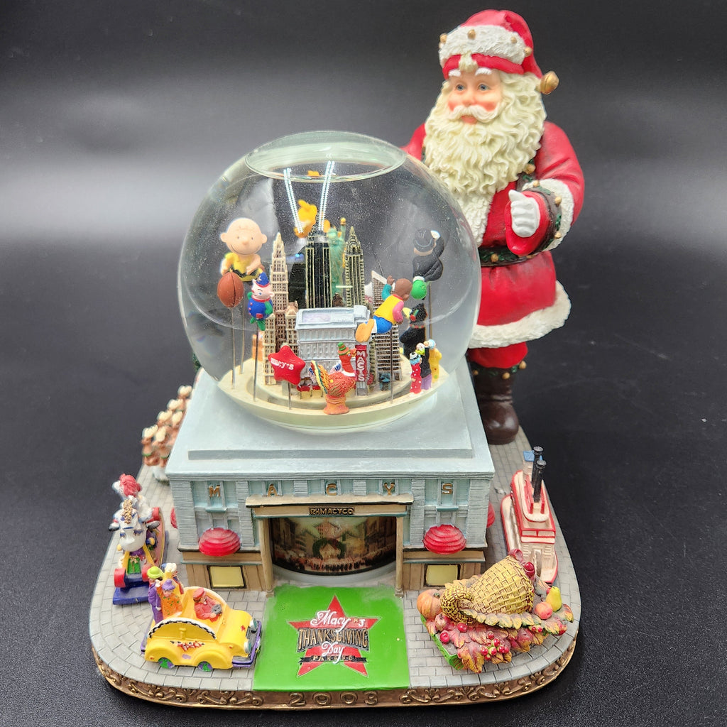A festive Santa Claus snow globe music box featuring a large clear glass dome with miniature holiday figures and snow, set on a detailed base with Santa standing behind it, against a dark gray background. Santa snowglobe Macy's Day Parade 2003
