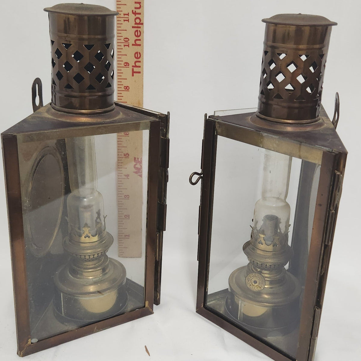 Two vintage oil lanterns with dark bronze metal frames and clear glass panels, shown side by side with a wooden ruler for scale against a white background.