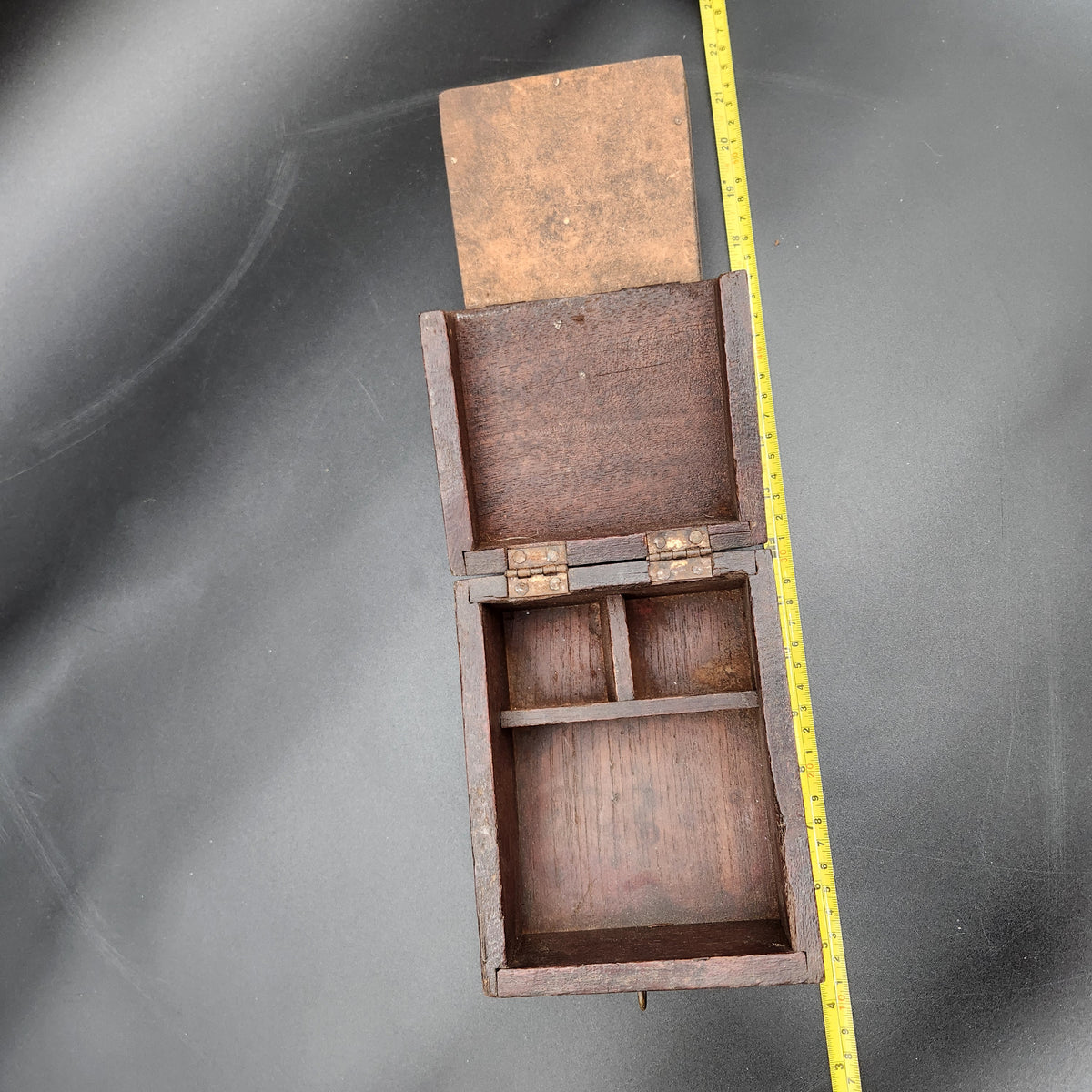 A small vintage wooden box with an open hinged lid and internal compartments, shown on a dark gray background next to a yellow measuring tape for scale.