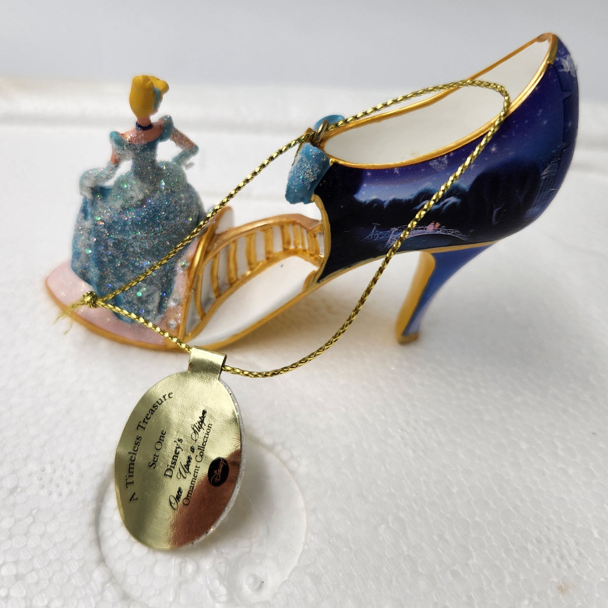 A miniature Cinderella-themed necklace featuring a blue high-heeled shoe with a painted landscape, a small Cinderella figurine, and a gold-tone oval pendant with engraved text, displayed on a white textured surface.