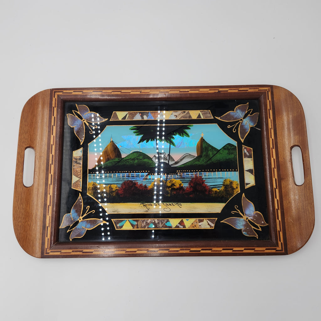 A rectangular wooden serving tray with rounded corners and integrated side handles, featuring a glossy black center panel decorated with a colorful landscape scene and butterfly motifs. Vintage Brazilian Butterfly Wing Marquetry Tray Rio De Janeiro Inlaid Wood Label