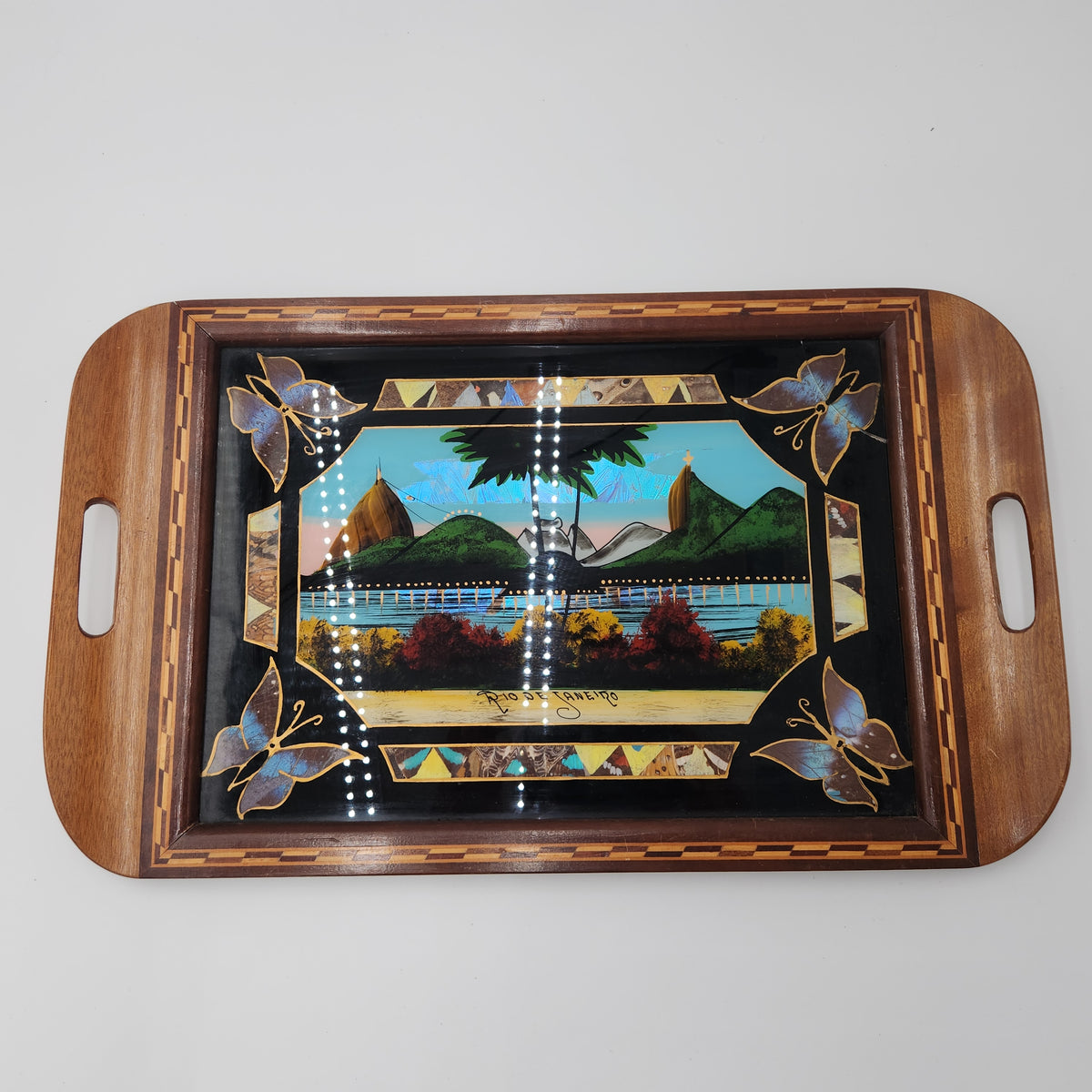 A rectangular wooden serving tray with rounded corners and integrated side handles, featuring a glossy black center panel decorated with a colorful landscape scene and butterfly motifs. Vintage Brazilian Butterfly Wing Marquetry Tray Rio De Janeiro Inlaid Wood Label