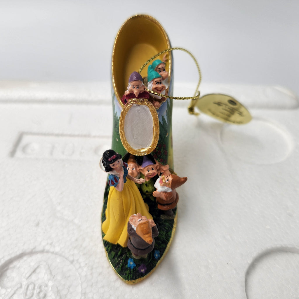 A Disney-themed ornament shaped like a high-heeled shoe, featuring Snow White, the Seven Dwarves, and a small oval mirror, displayed on a white foam packaging surface.
