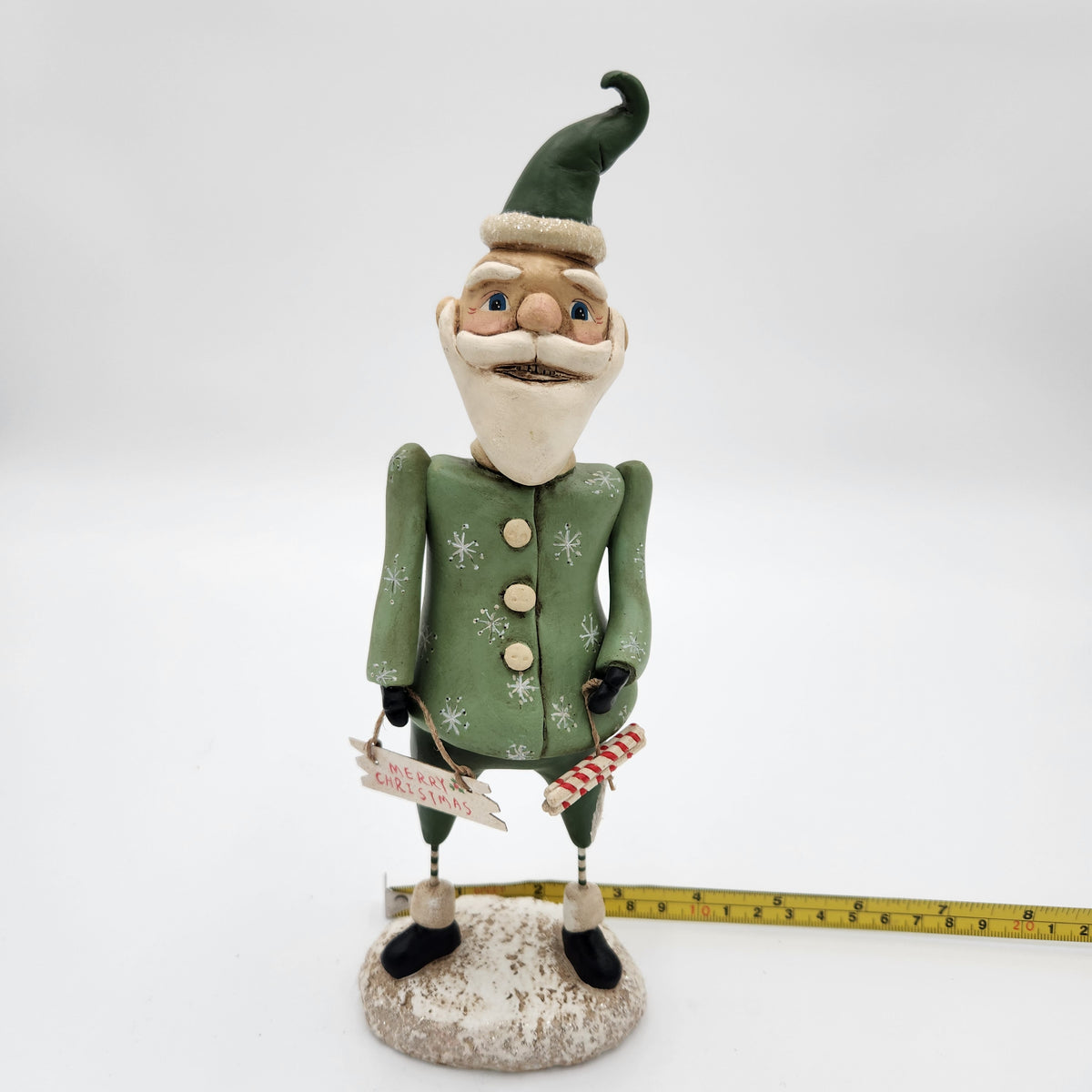 A whimsical Santa Claus figurine dressed in a green elf-style coat with white snowflake patterns, holding a candy cane and a small sign, standing on a textured base with a yellow measuring tape placed behind it on a white background. Janell Berryman Pumpkinseeds "Sage and Peppermint Santa