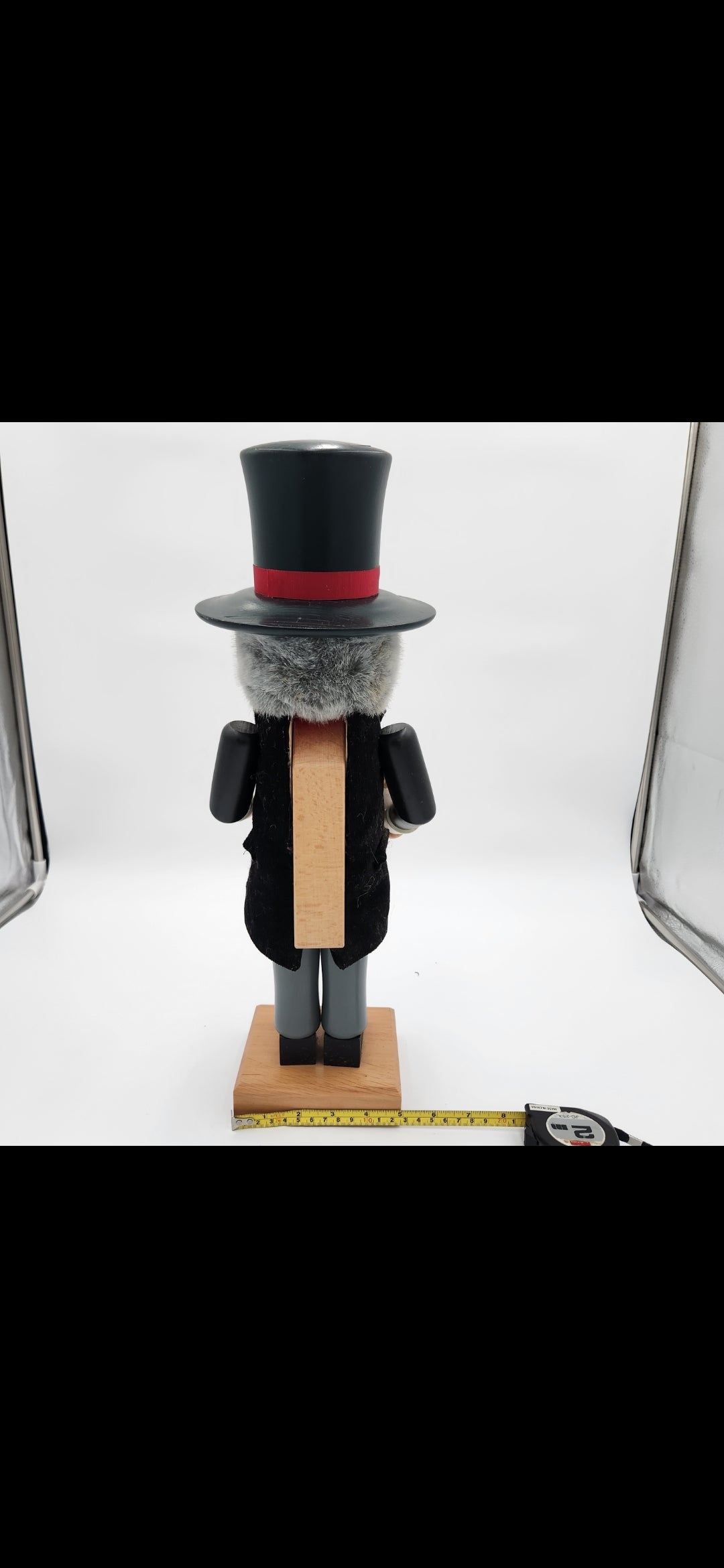 A vintage wooden nutcracker figurine shown from the back, wearing a black top hat with a red band, a black coat, gray legs, and standing on a wooden base. A tape measure is placed in front of the base to show its height.Christian Ulbrickt