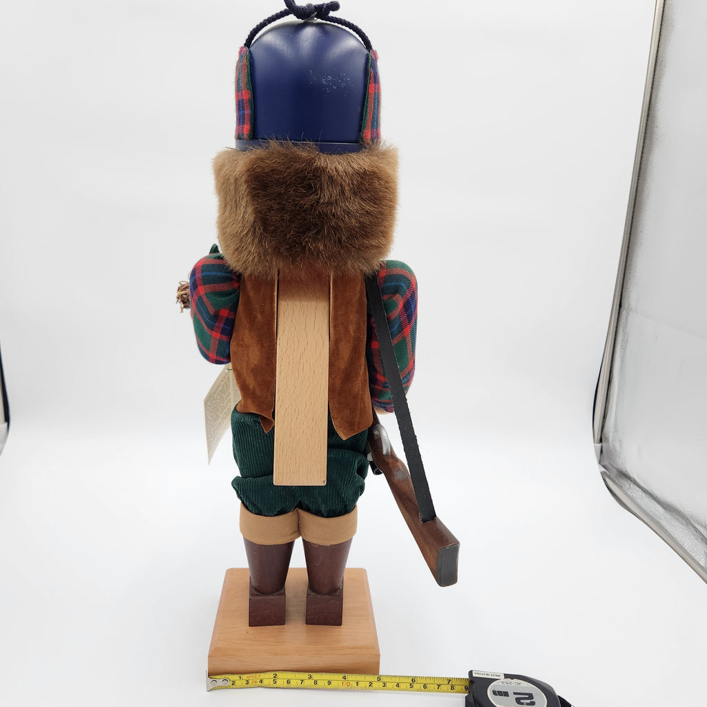 A rustic wooden  Christian Ulbricht Nutcracker The Duck Hunter shown from the back, featuring a dark blue hat with red plaid trim, brown faux fur, a brown suede vest, green pants, and a wooden base. A tape measure is visible at the bottom to show the height, and a studio light reflector is on the right side.
