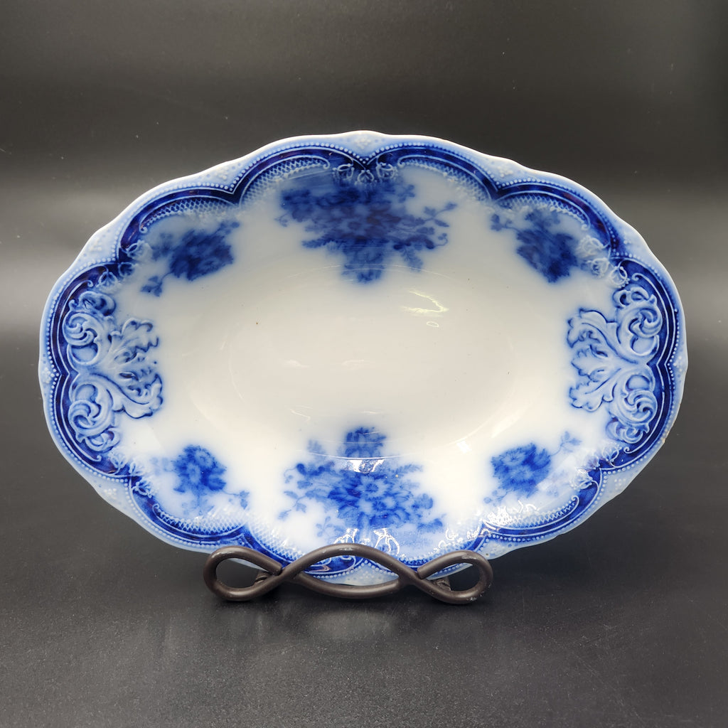 An oval white porcelain serving bowl with blue floral and scroll patterns, displayed on a dark metal stand against a dark gray background. Antique 1890s Flow Blue Cobalt Oval Serving Bowl Deep Vegetable Dish