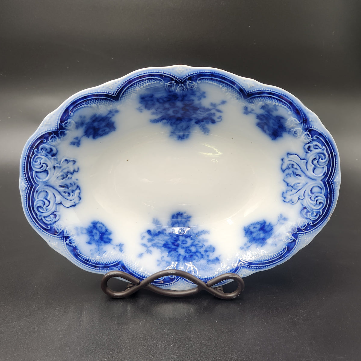 An oval white porcelain serving bowl with blue floral and scroll patterns, displayed on a dark metal stand against a dark gray background. Antique 1890s Flow Blue Cobalt Oval Serving Bowl Deep Vegetable Dish