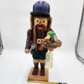 A traditional handcrafted wooden  Christian Ulbricht Nutcracker The Duck Hunter figurine of a bearded hunter wearing a blue bowler hat, red plaid shirt, and green shorts, holding a duck in a nest and a pistol. The figurine stands on a wooden base with a measuring tape visible at the bottom and a product tag hanging from its belt.