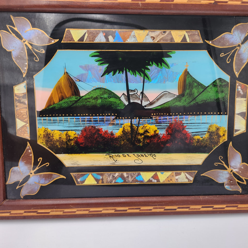 A framed decorative art piece featuring a colorful scenic illustration with green hills, a pier, palm trees, and a central black silhouette. The frame has butterfly motifs in the corners and geometric border details. Vintage Brazilian Butterfly Wing Marquetry Tray Rio De Janeiro Inlaid Wood Label