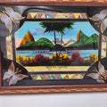 A framed decorative art piece featuring a colorful scenic illustration with green hills, a pier, palm trees, and a central black silhouette. The frame has butterfly motifs in the corners and geometric border details. Vintage Brazilian Butterfly Wing Marquetry Tray Rio De Janeiro Inlaid Wood Label