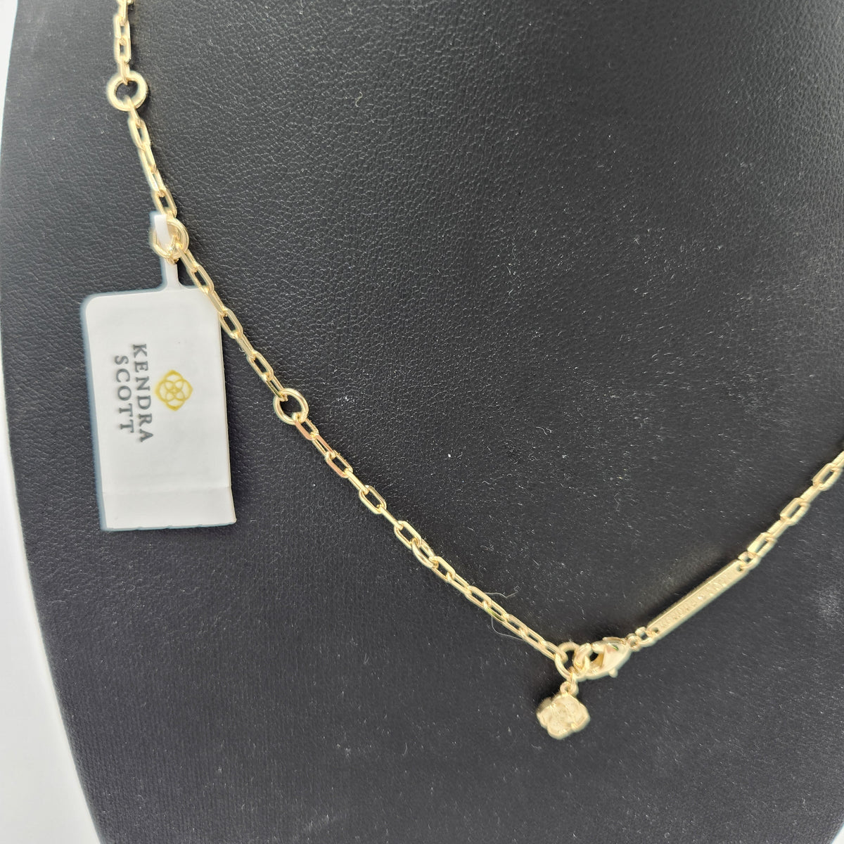 A  Kendra Scott gold chain necklace with a small textured cube charm, displayed on a black jewelry bust next to a Kendra Scott tag.