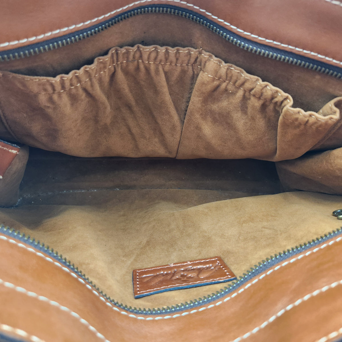 Close-up of the interior of a Patricia Nash brown leather bag, showing a tan suede-like lining, a zippered compartment, and a small brown leather brand patch near the bottom.