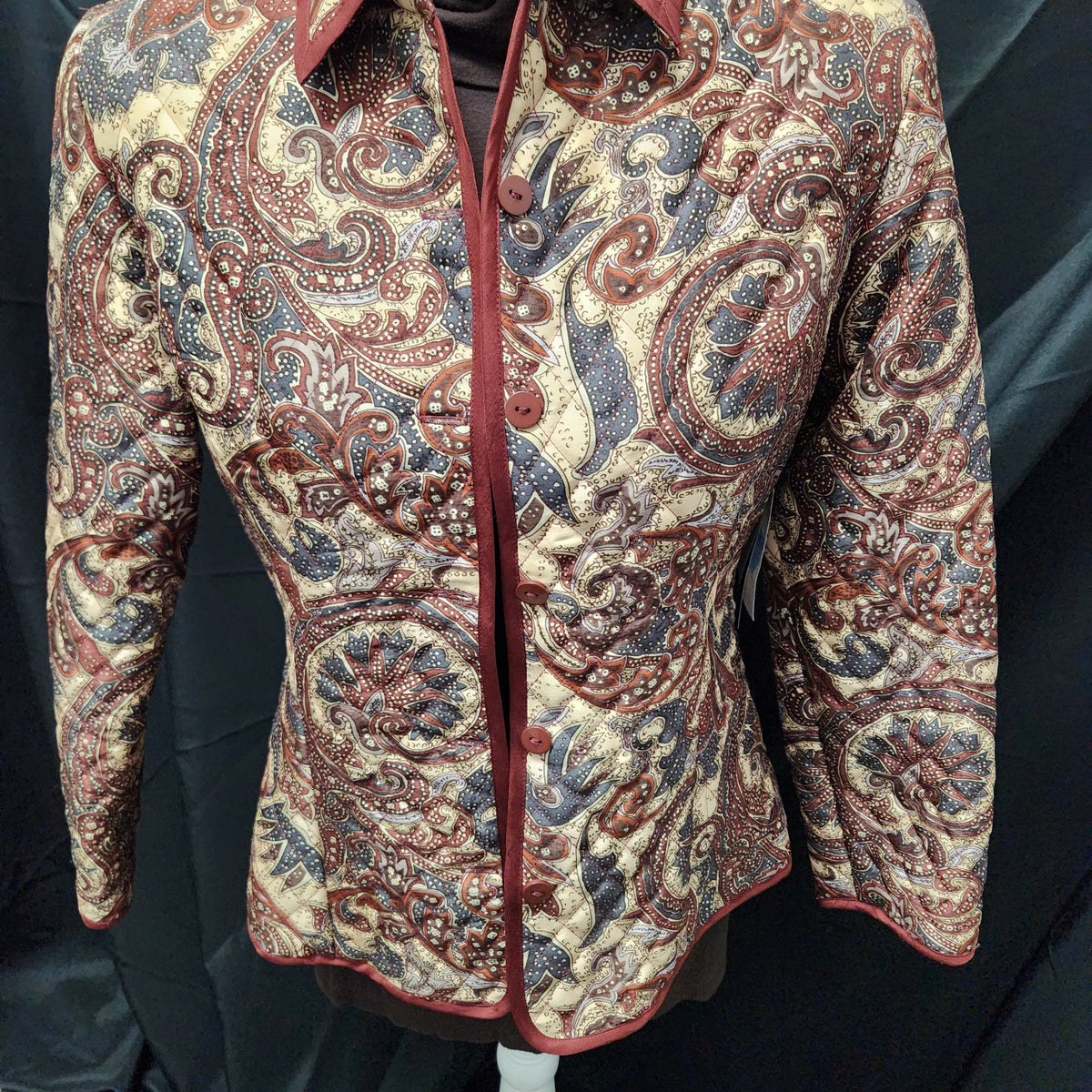 A long-sleeve button-front shirt jacket with an intricate paisley pattern in beige, rust red, and blue, displayed on a mannequin against a black fabric backdrop.