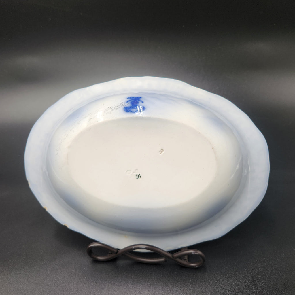 An oval white ceramic plate or dish with a blue painted motif near the rim, displayed upside down on a small black stand against a dark gray background. Antique 1890s Flow Blue Cobalt Oval Serving Bowl Deep Vegetable Dish