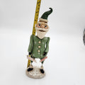 A handcrafted Christmas elf figurine standing on a textured base, holding a sign and candy cane, with a yellow tape measure behind it for scale, on a white background. Janell Berryman Pumpkinseeds "Sage and Peppermint Santa
