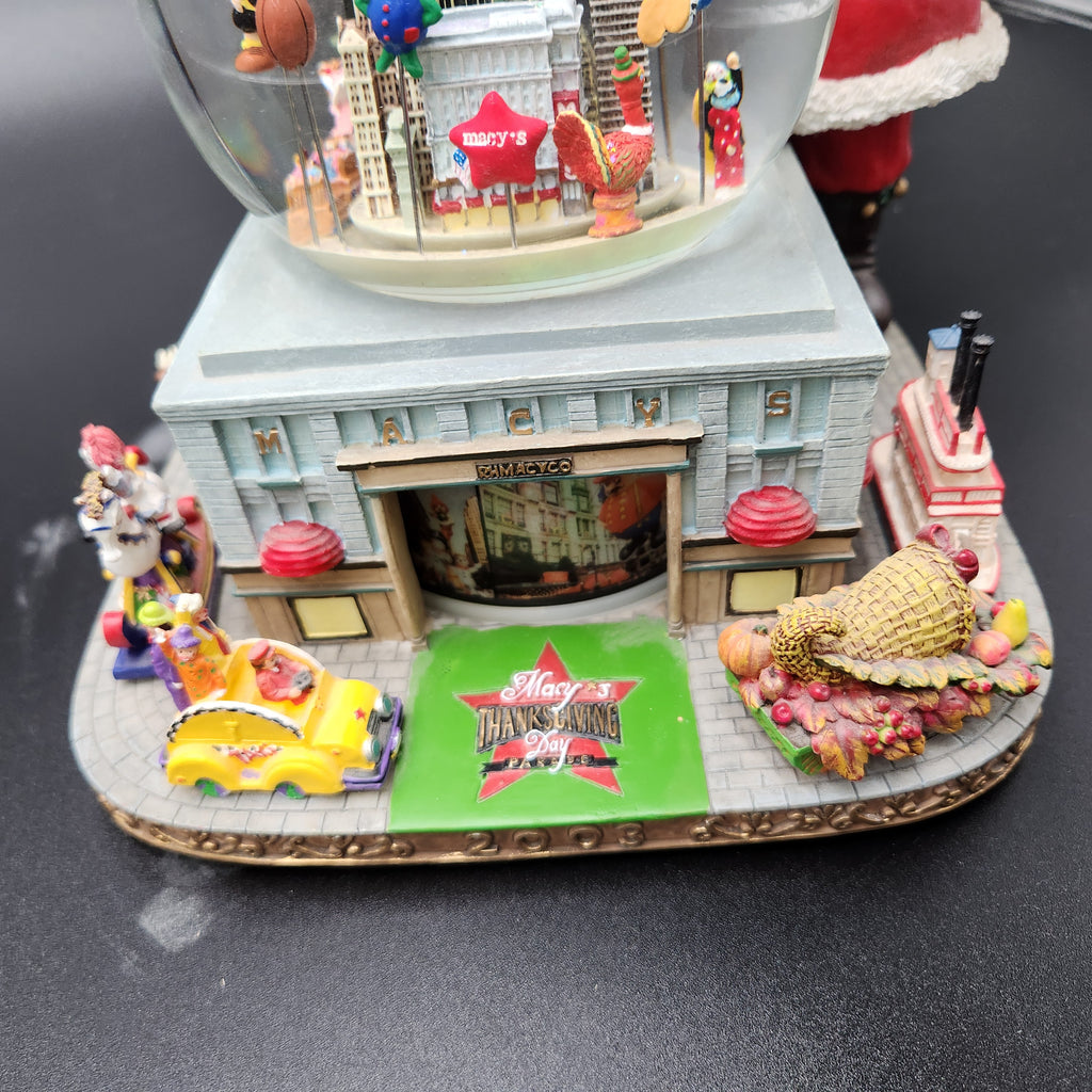 A detailed miniature Thanksgiving-themed snow globe base featuring a gray building with a storefront, a yellow car, a cornucopia with pumpkins, and a green sign reading 'Merry Thanksgiving.' The clear globe at the top contains tiny figurines and city-like decorations. Santa snowglobe Macy's Day Parade 2003