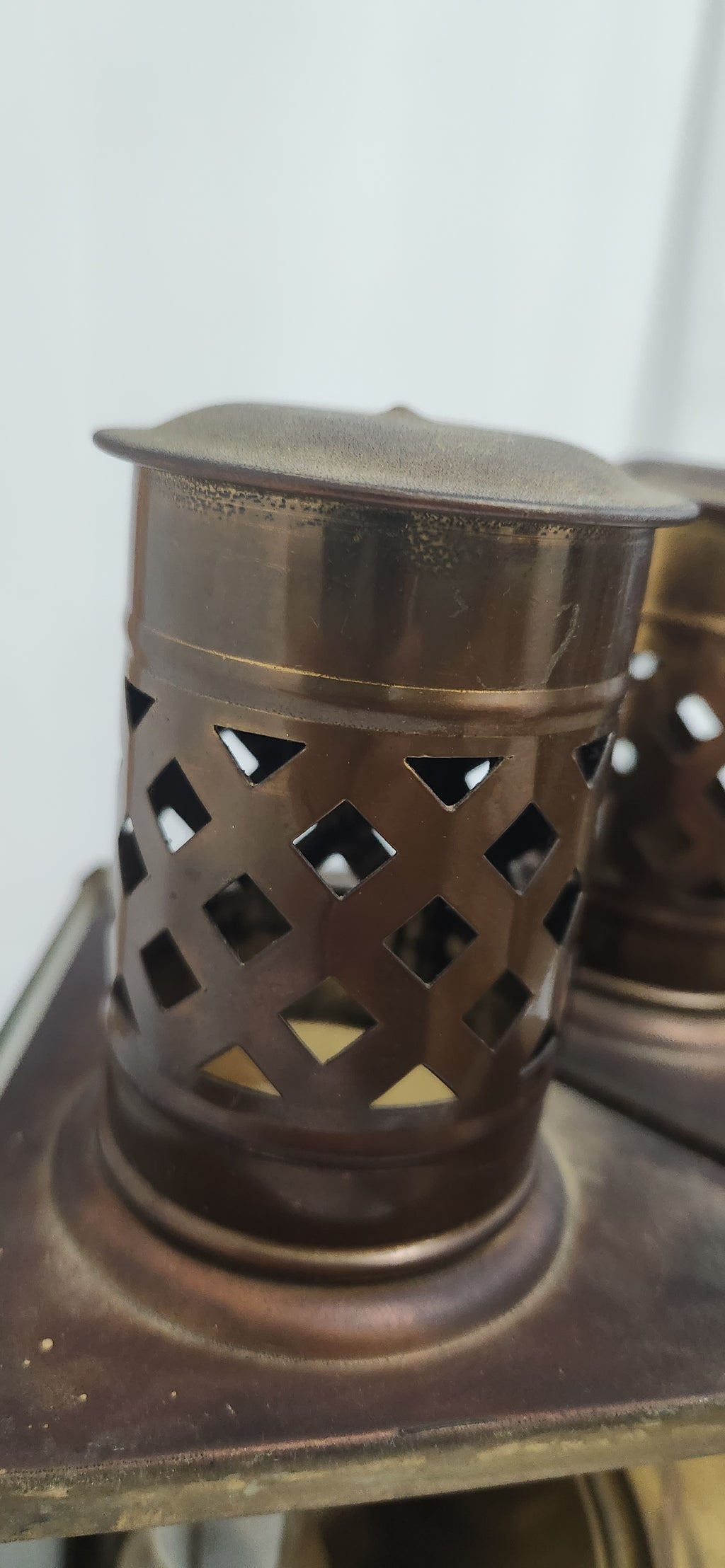 A close-up of a small cylindrical metal lantern or candle holder with a perforated diamond lattice pattern on the sides, shown in a warm bronze or copper-toned finish.