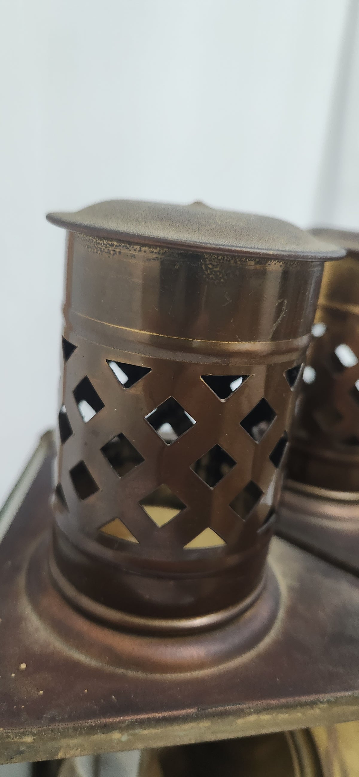 A close-up of a small cylindrical metal lantern or candle holder with a perforated diamond lattice pattern on the sides, shown in a warm bronze or copper-toned finish.