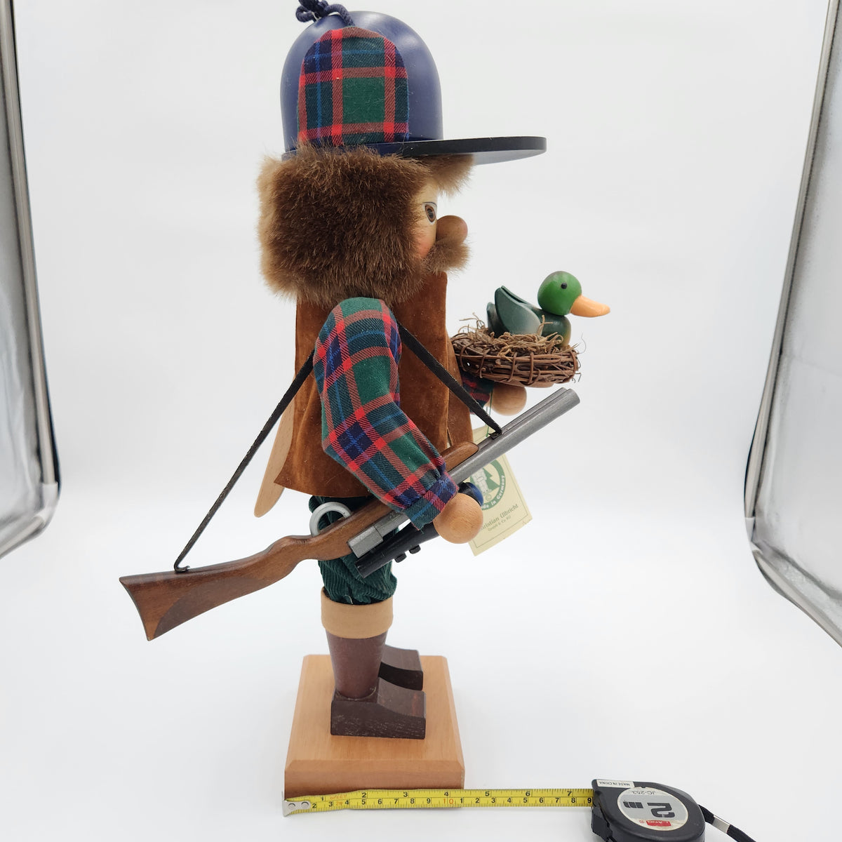 A wooden  Christian Ulbricht Nutcracker The Duck Hunter on a white studio background, shown with a measuring tape at its base. The figure features a brown fur-trimmed hat, a red and green plaid shirt and hat, a brown beard, a rifle, and holds a small green duck in a nest.