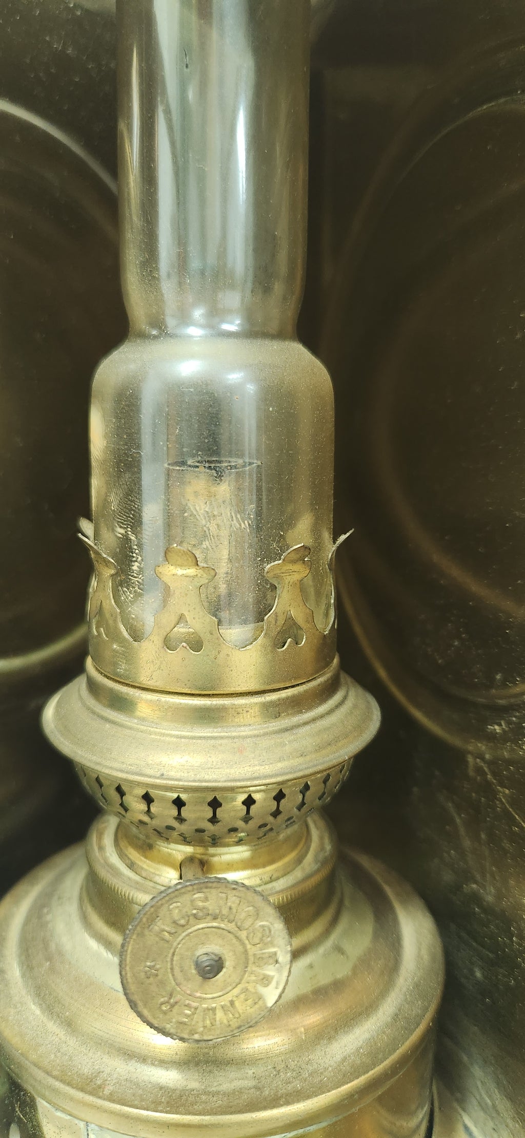 Close-up of a vintage brass oil lamp with a clear glass chimney and decorative cutout details, showing a circular brass dial on the base.