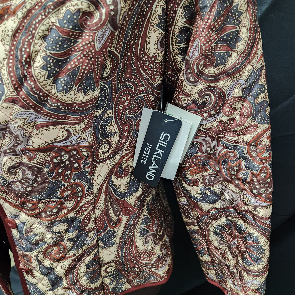 A close-up of a patterned silkland jacket with a paisley design in beige, red, and navy blue, featuring a black Silkland Boutique brand tag.
