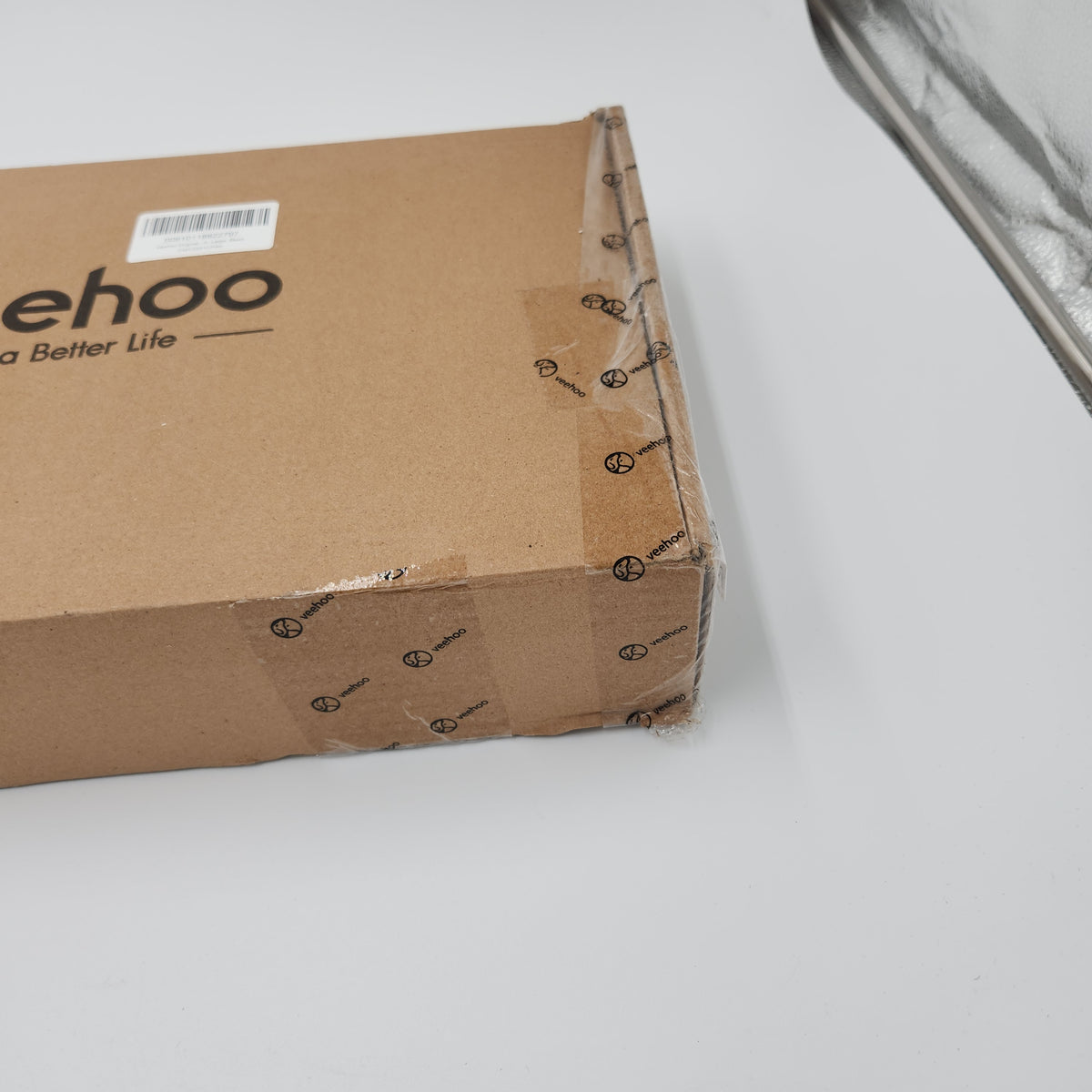 A close-up of a brown cardboard box sealed with clear tape, showing partial brand text 'ehoo' on the side, photographed on a white surface.Veehoo elevated dog bed