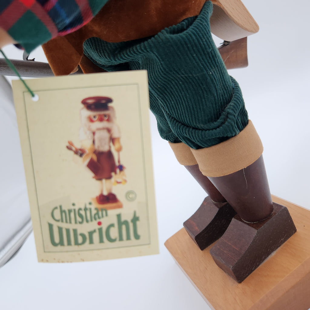 A close-up of  Christian Ulbricht Nutcracker The Duck Hunter showing its dark green ribbed tunic, brown boots, and wooden base, with a paper hangtag reading 'Christian Ulbricht' in the foreground.