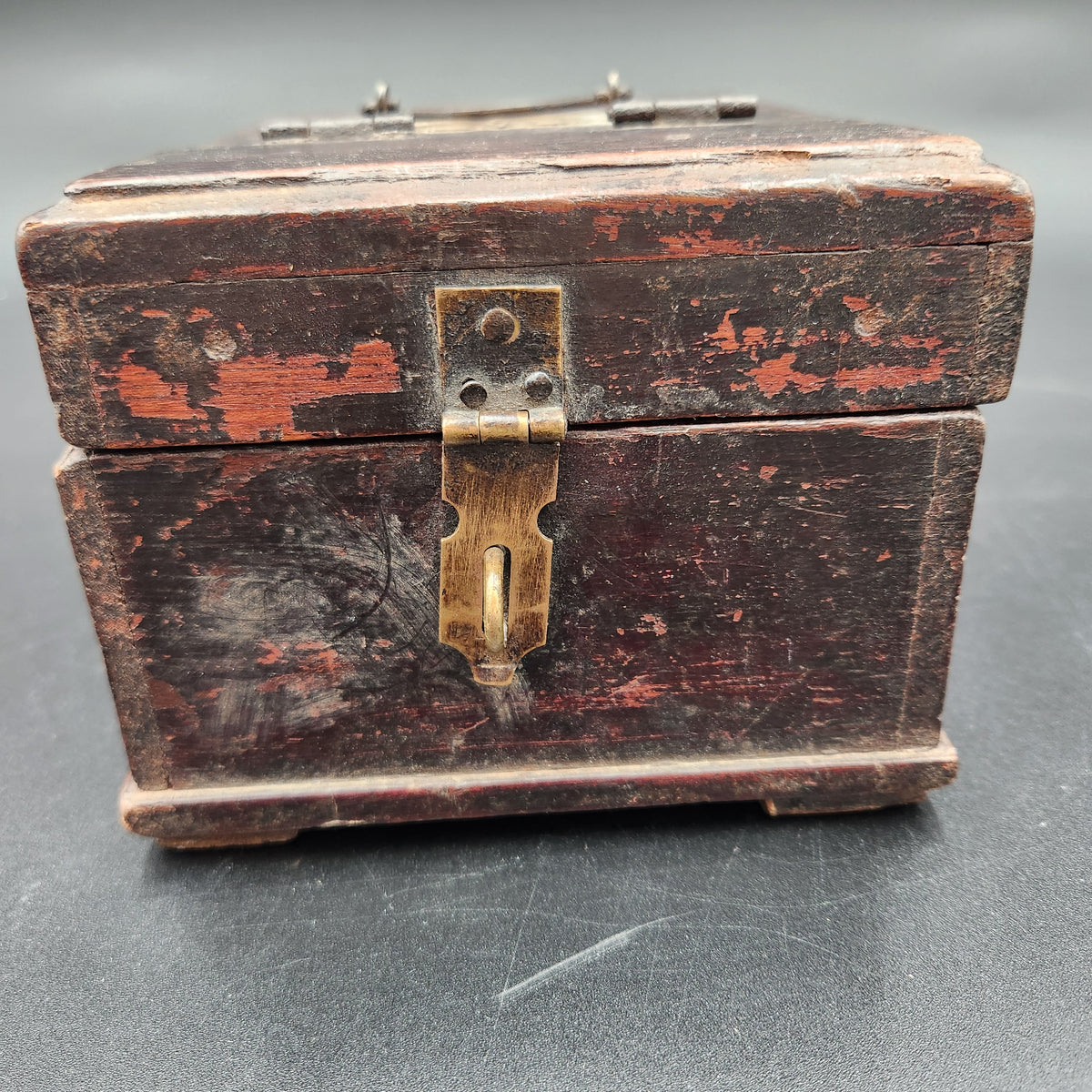 A small vintage wooden chest or jewelry box with a distressed reddish-brown finish and an antique brass latch on a dark gray background.